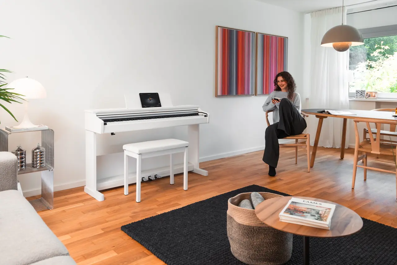 Weißes Kawai Digitalpiano in einem hellen Wohnzimmer; im Hintergrund sitzt eine Frau am Tisch.
